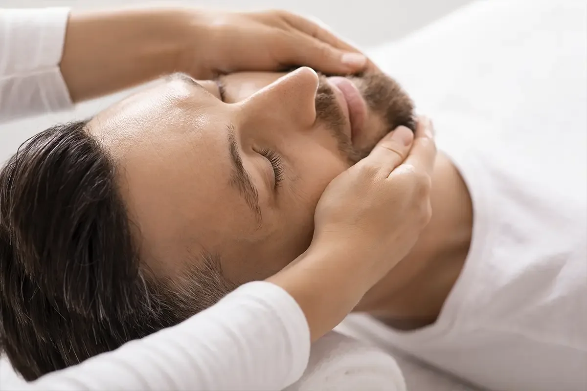 A practitioner&rsquo;s hands cradling a reclining man&rsquo;s head and jaw